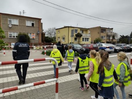 Zdjęcie przedstawia dzieci przechodzące przez przejście dla pieszych z policjantem i strażnikiem miejskim
