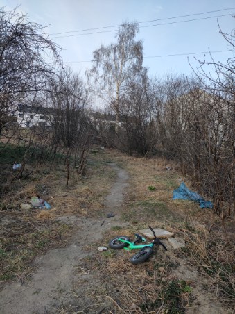 Zdjęcie przedstawia niezagospodarowany teren z trawą, drzewami i ścieżką, przy której leżą śmieci