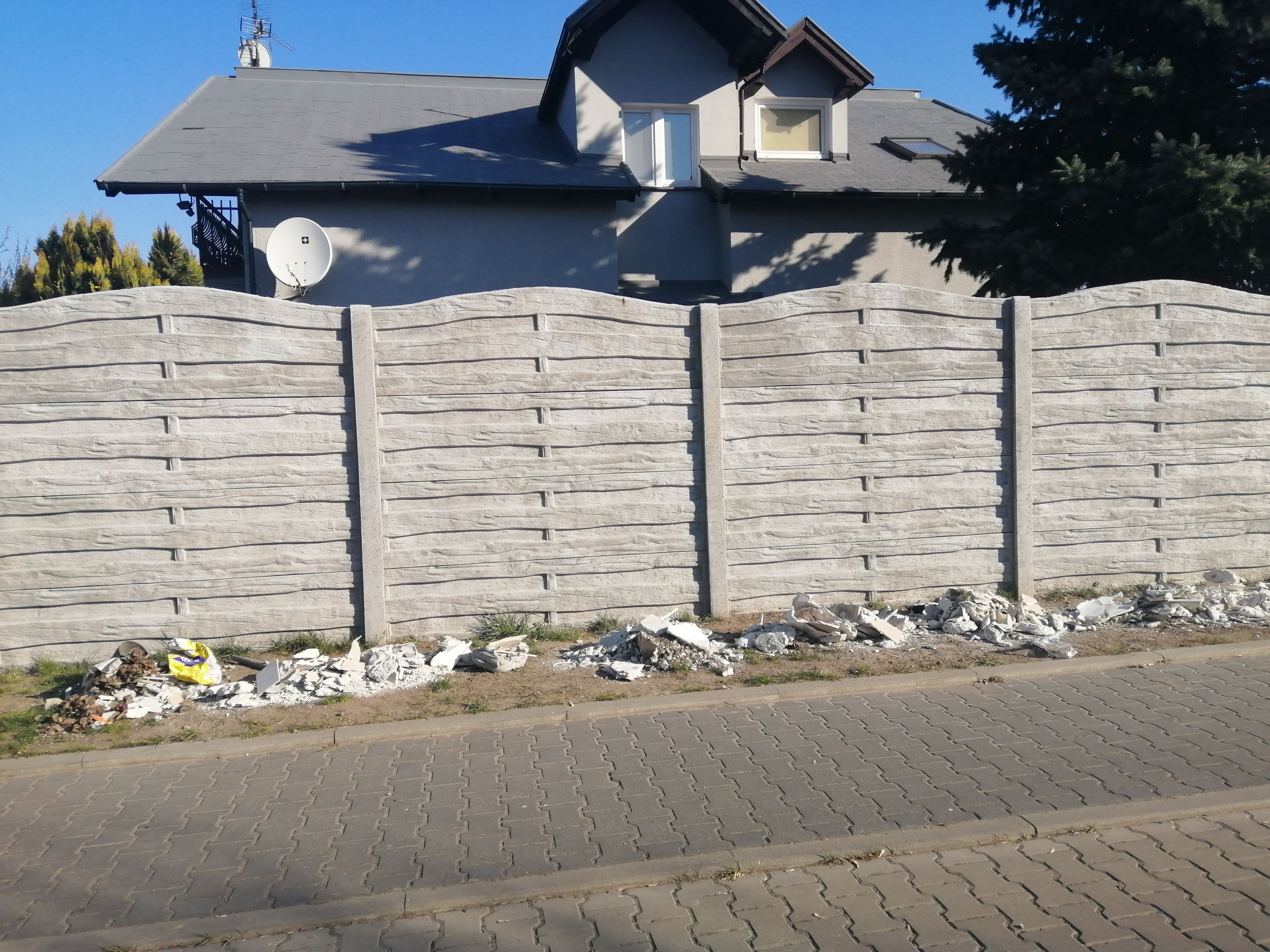 Zdjęcie przedstawia część ulicy oraz betonowy płot przy którym leżą odpady budowlane
