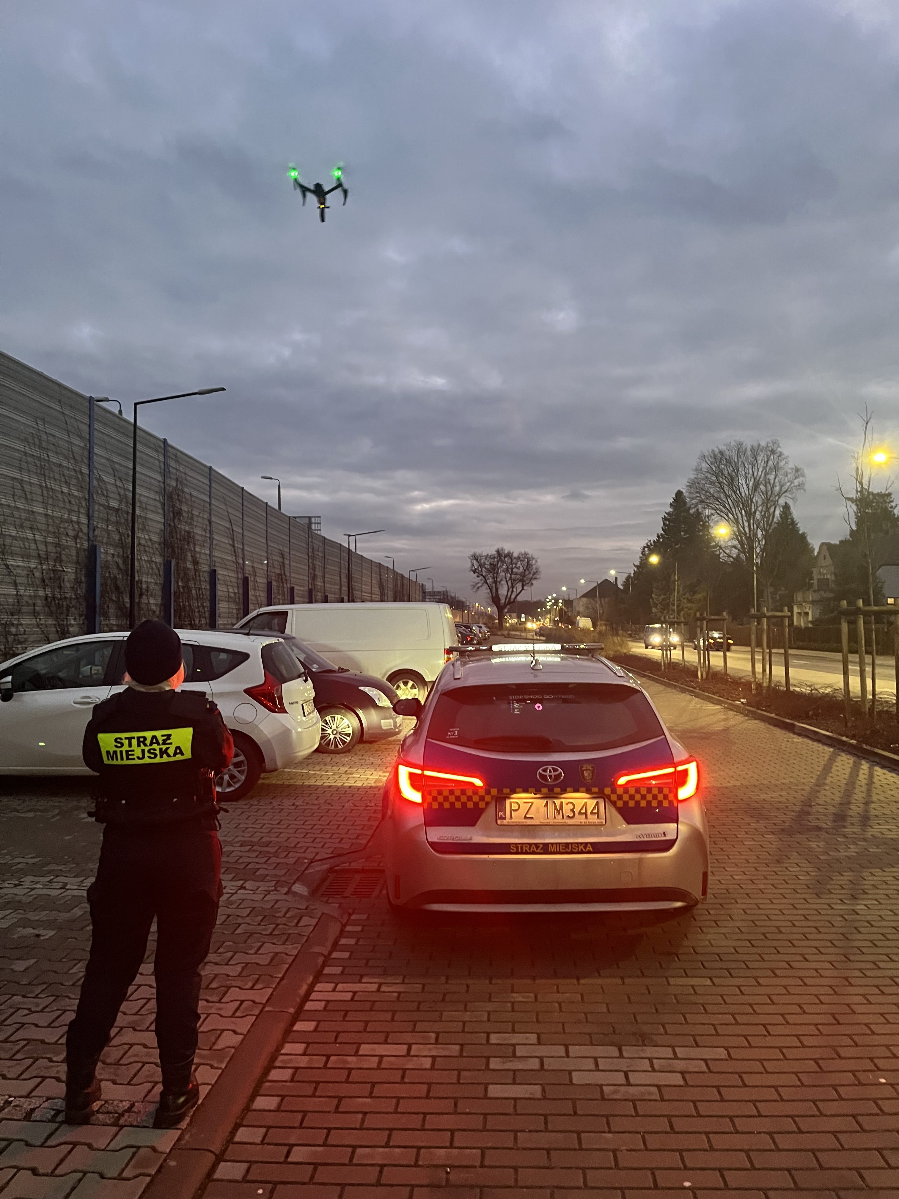 Zdjęcie przedstawia strażnika miejskiego przy radiowozie oraz wzbijający się w powietrze dron