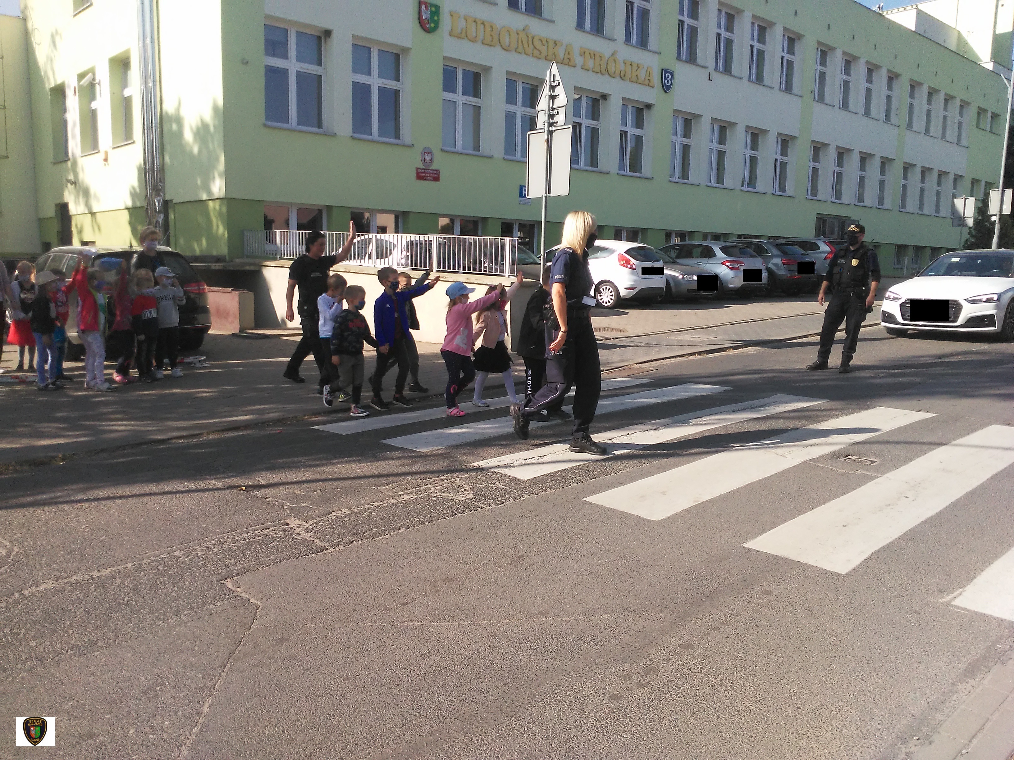 zdjęcie przedstawia dzieci przechodzące przez przejście dla pieszych w obecności funkcjonariuszy straży miejskiej i policji