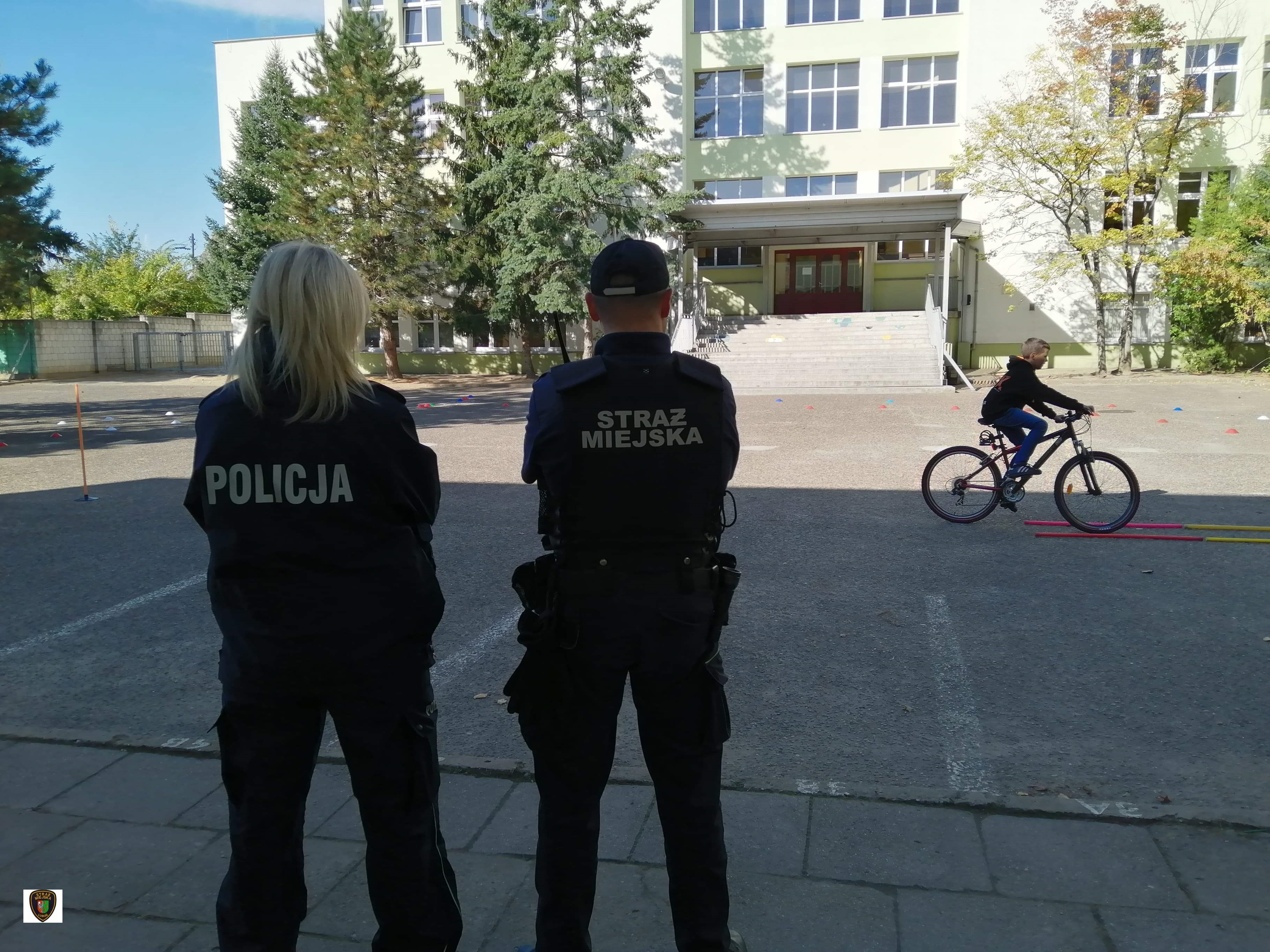 Zdjęcie przedstawia stojących policjantkę i strażnika miejskiego oraz dziecko jadące rowerem podczas egzaminu na kartę rowerową