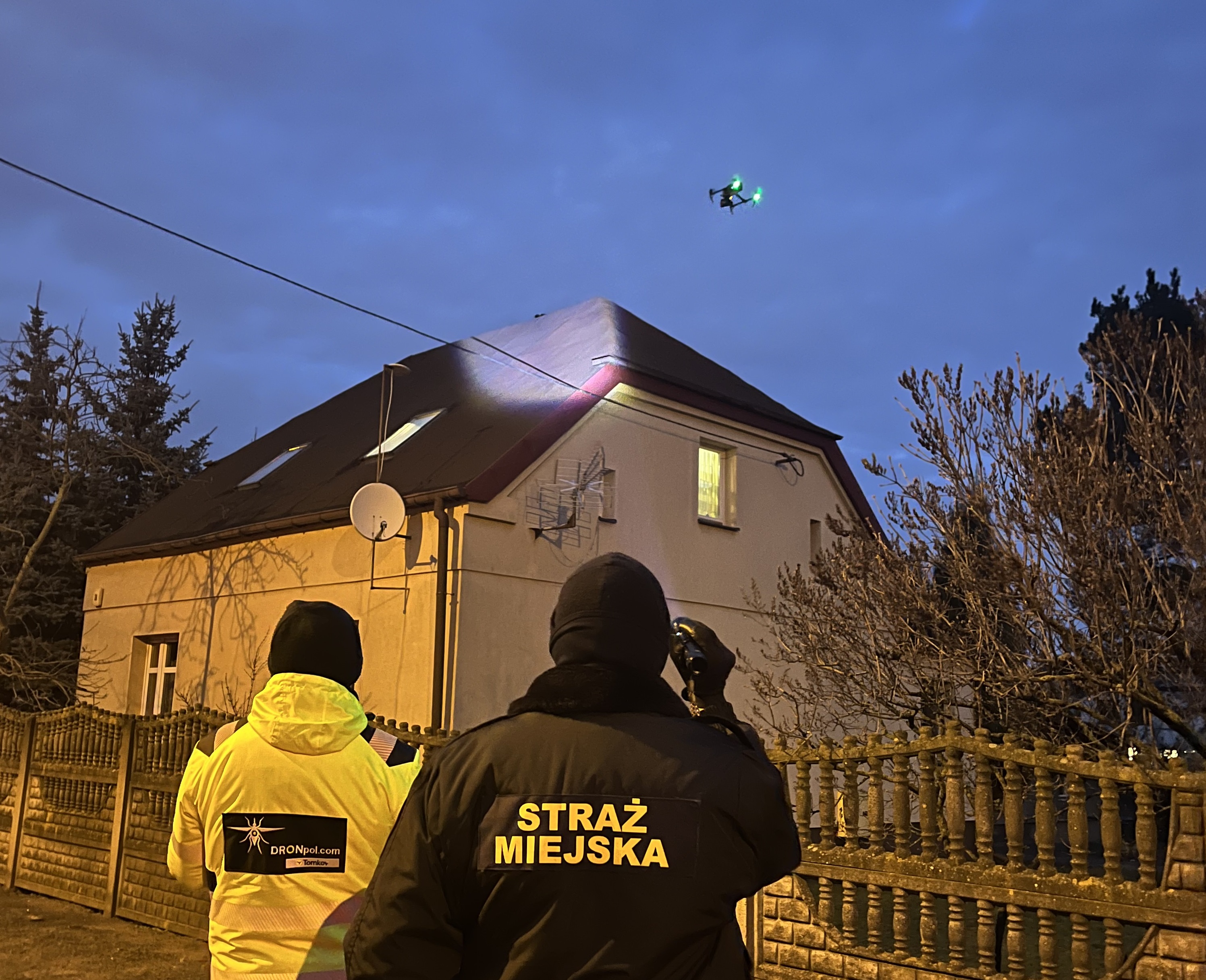 Zdjęcie przedstawia strażnika miejskiego, osobę obsługującą drona oraz drona unoszącego się nad dachem domu